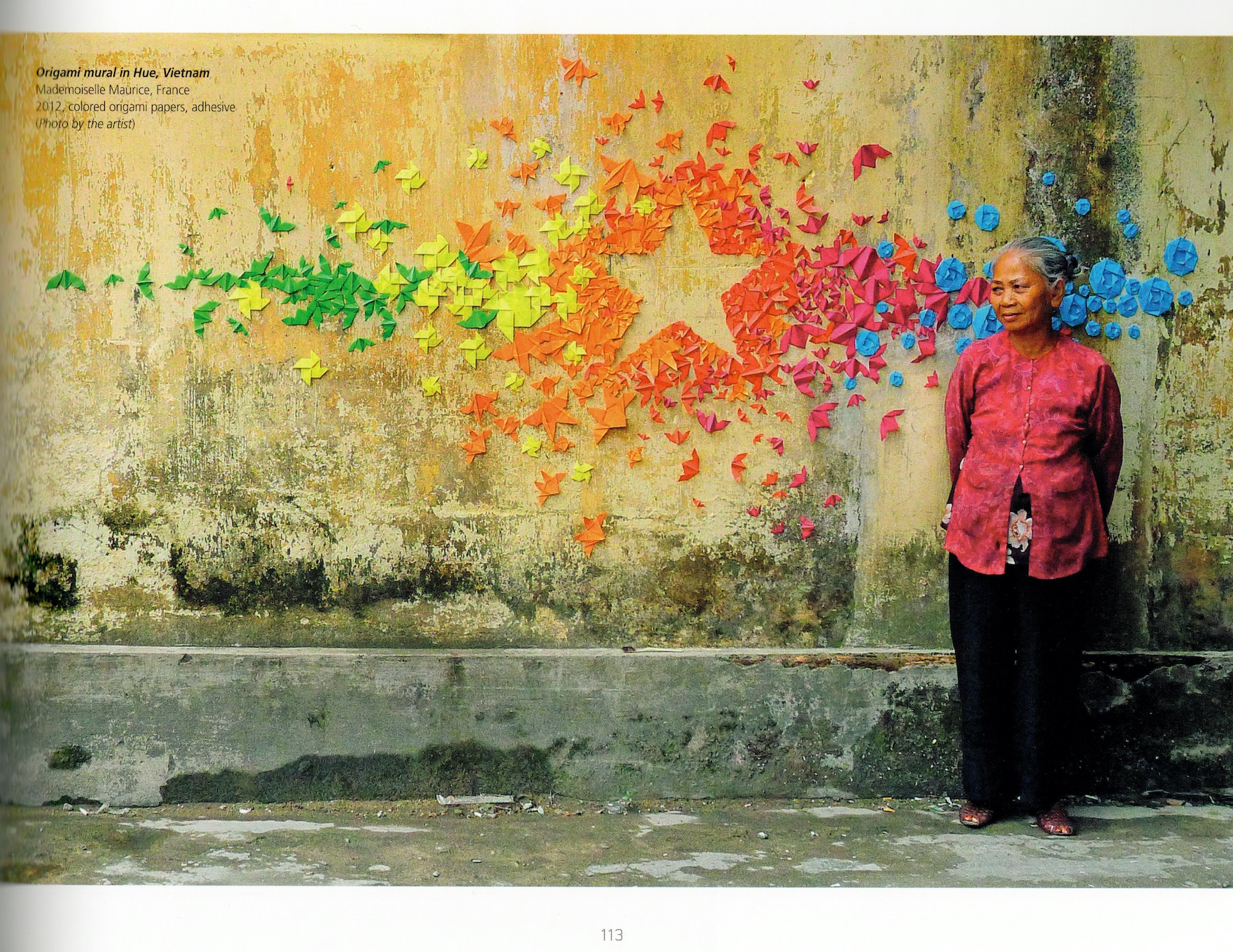 Older woman in pink and black stands in front of a stone wall with various rainbow origami pieces together that collectively create the borders of a star