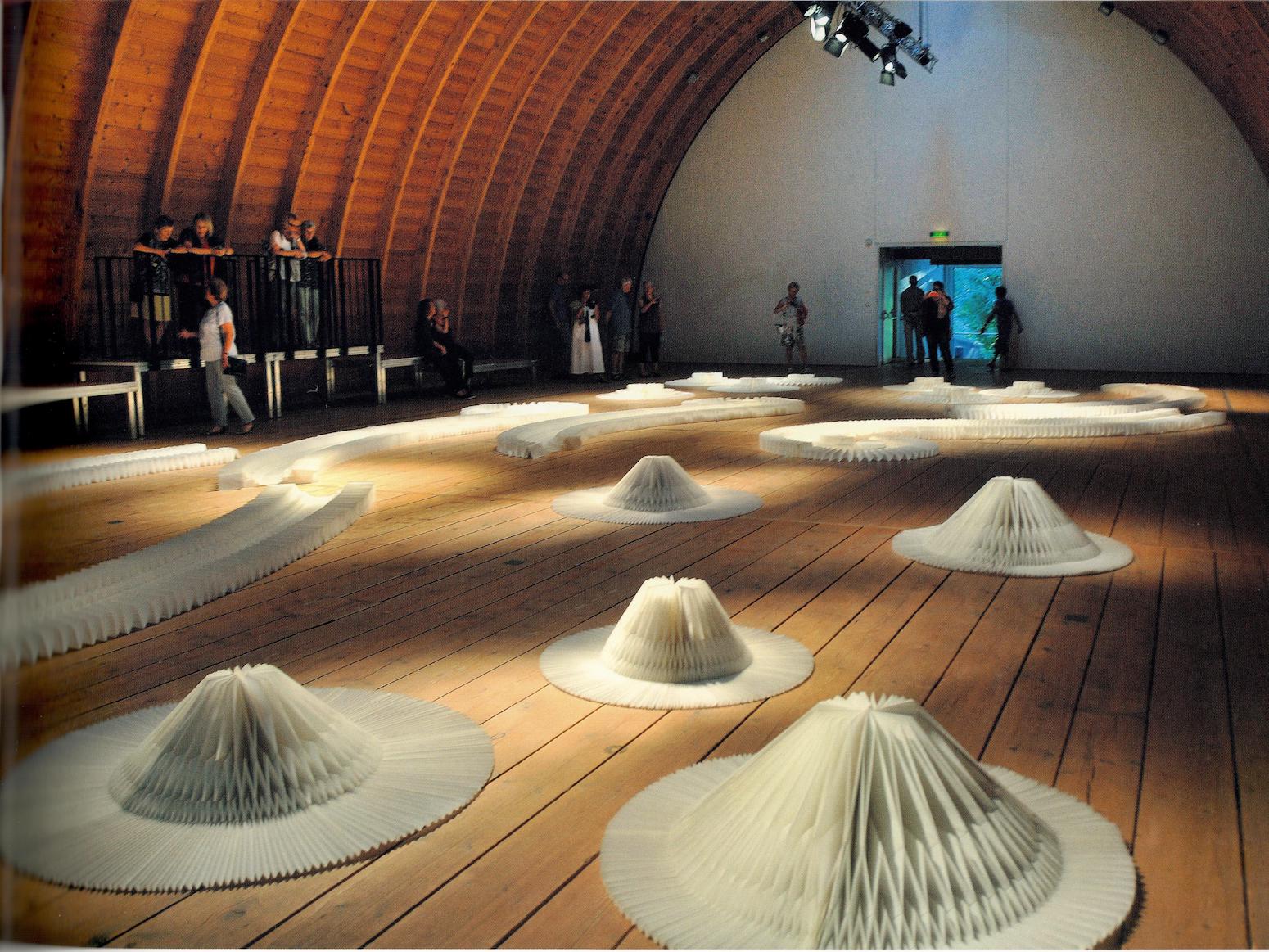 Wooden room with curved ceiling, with over a dozen largescale white origami sculptures scattered around the room